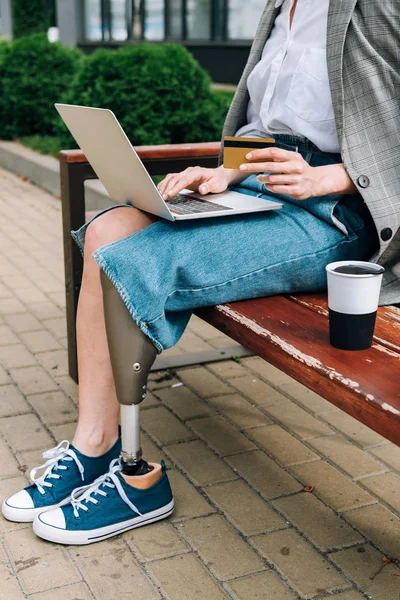 Vue partielle de la femme handicapée avec ordinateur portable assis sur le banc et tenant la carte de crédit — Photo de stock