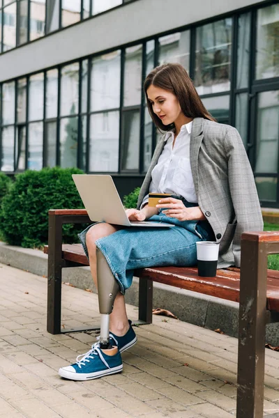 Femme handicapée avec ordinateur portable assis sur le banc et tenant la carte de crédit — Photo de stock