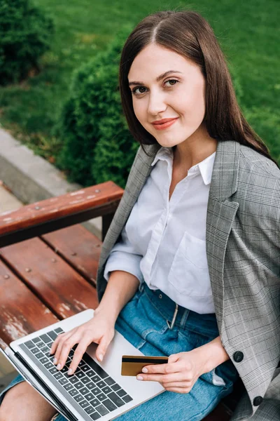 Sonriente mujer joven con el ordenador portátil sentado en el banco y la celebración de la tarjeta de crédito - foto de stock