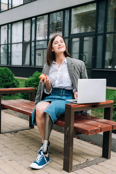 Улыбающаяся женщина-инвалид с яблоком сидит на скамейке и использует ноутбук на улице — стоковое фото