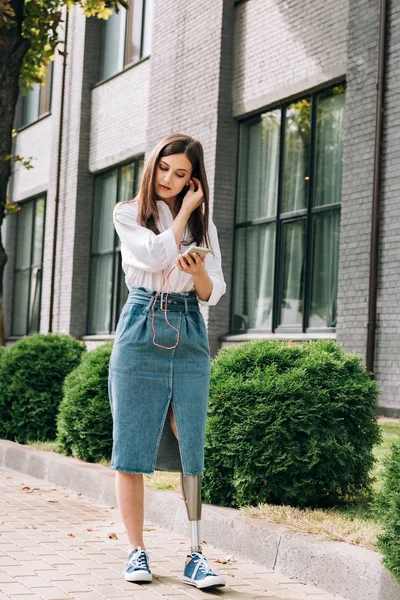 Vista integrale della donna disabile che tiene lo smartphone con gli auricolari sulla strada — Foto stock