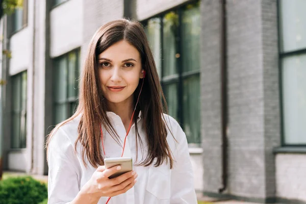 Sorrindo jovem mulher em camisa branca segurando smartphone e ouvindo música em fones de ouvido na rua — Fotografia de Stock