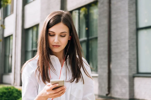 Attrayant jeune femme en chemise blanche tenant smartphone et écouter de la musique dans les écouteurs sur la rue — Photo de stock