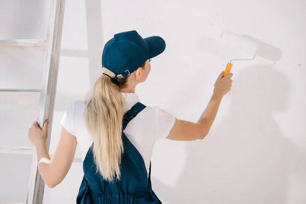 Вид на молодого художника в форме, стоящего у белой стены и держащего в руках ролик с краской — стоковое фото