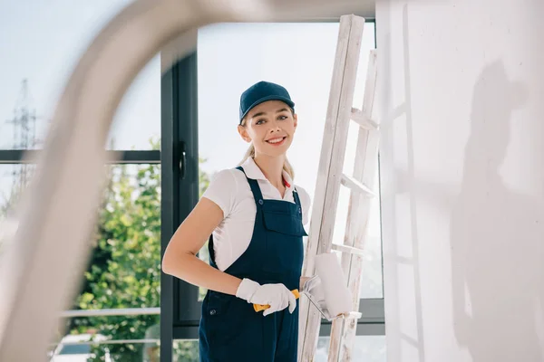 Foco seletivo de pintor muito jovem em uniforme segurando rolo de pintura e sorrindo para a câmera — Fotografia de Stock