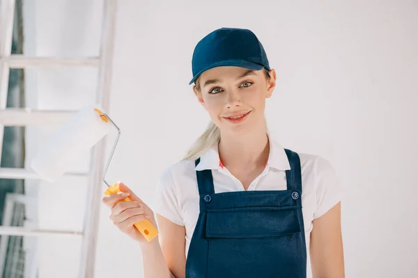 Belle jeune femme en uniforme tenant rouleau de peinture et souriant à la caméra — Photo de stock