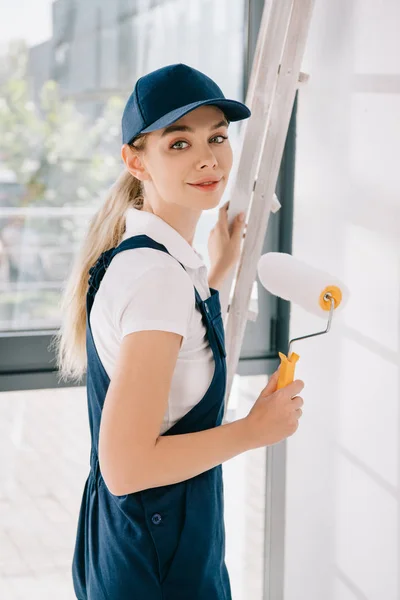 Pretty young painter in uniform holding paint roller and smiling at camera — Stock Photo