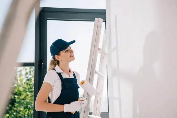 Вибірковий фокус молодого художника в уніформі, що стоїть біля сходів, тримає фарбувальний валик і дивиться на білу стіну — стокове фото