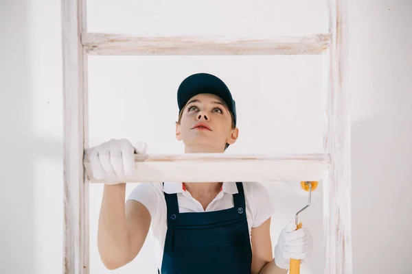 Bastante joven pintor de uniforme mirando hacia arriba mientras sube en la escalera - foto de stock