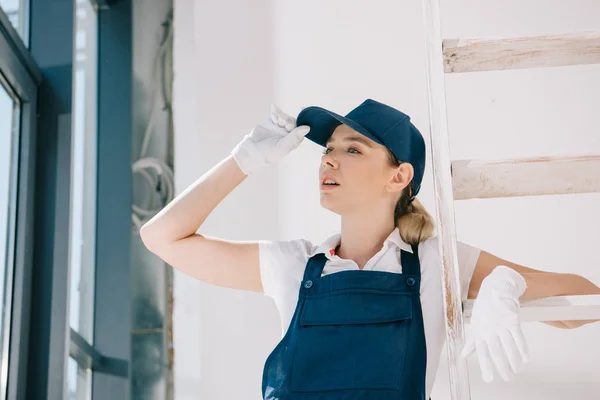 Jolie jeune peintre en uniforme toucher cap tout en regardant loin — Photo de stock