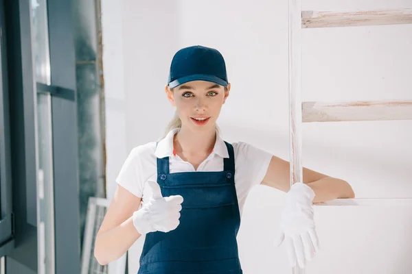 Bastante joven pintor en uniforme mostrando el pulgar hacia arriba mientras mira a la cámara - foto de stock