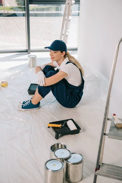 Beautiful young painter in overalls and cap sitting on floor near paint roller in roller tray and cans with paint — Stock Photo