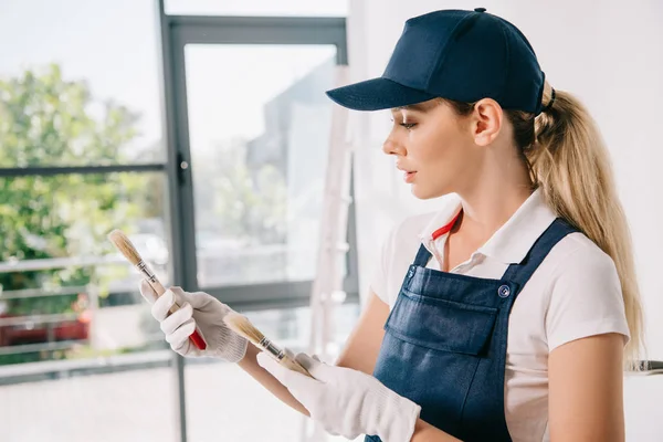 Beautiful young painter in overalls and cap holding paintbrushes — Stock Photo