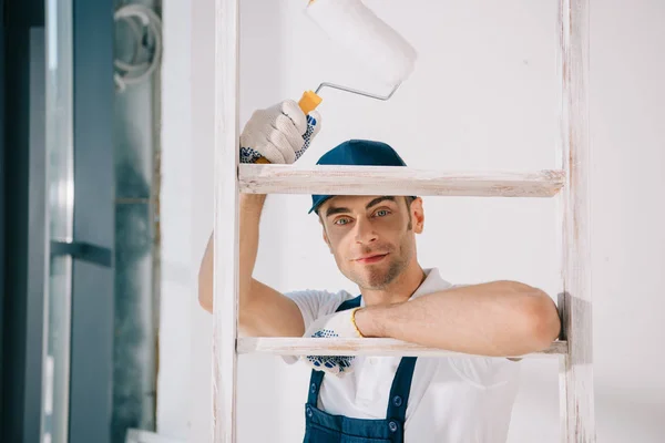 Beau jeune peintre en uniforme debout près de l'échelle, tenant rouleau de peinture et souriant à la caméra — Photo de stock