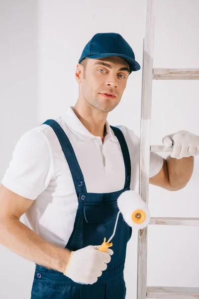 Bonito jovem pintor em uniforme de pé perto escada, segurando rolo de pintura e olhando para a câmera — Fotografia de Stock