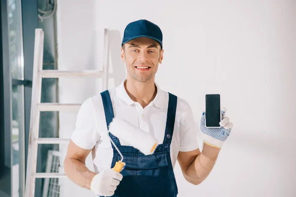 Joven pintor guapo en uniforme sosteniendo rodillo de pintura y mostrando teléfono inteligente con pantalla en blanco — Stock Photo