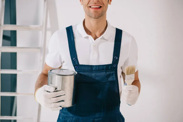 Cropped view of smiling painter in uniform holding paintbrush and can with paint — Stock Photo