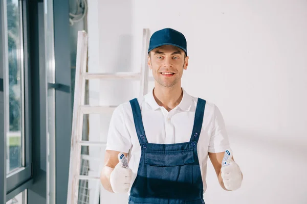 Schöner Maler in Uniform zeigt Daumen hoch und lächelt in die Kamera — Stockfoto