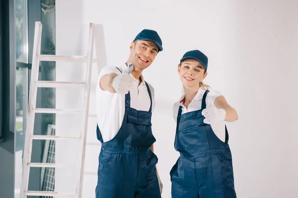 Young painters in overalls showing thumbs up while smiling at camera — Stock Photo