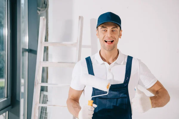 Joven pintor guapo en uniforme sosteniendo rodillo de pintura, mostrando el pulgar hacia arriba y sonriendo a la cámara - foto de stock