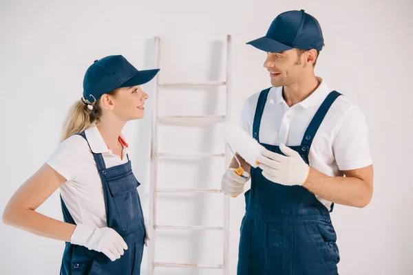 Beau peintre en uniforme tenant rouleau de peinture tout en parlant avec un collègue attrayant — Photo de stock