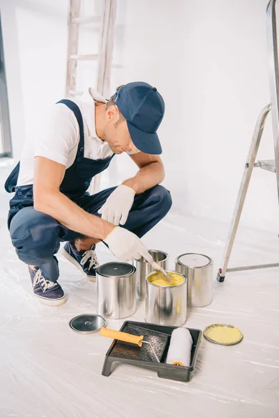 Junge Malerin in Overalls und Mütze holt mit Pinsel gelbe Farbe aus Dose — Stockfoto