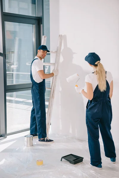 Bonito pintor de pé perto escada enquanto bonito colega segurando rolo de pintura — Fotografia de Stock