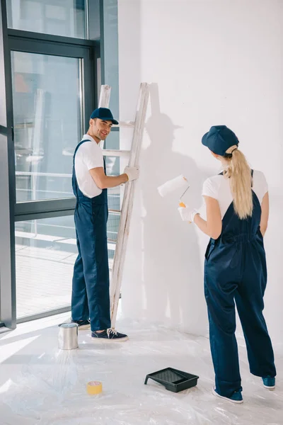 Pintor guapo de pie cerca de la escalera mientras bastante colega sosteniendo rodillo de pintura - foto de stock