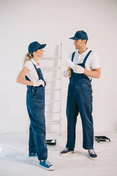 Pintor bonito em uniforme segurando rolo de pintura enquanto conversa com colega bonita — Fotografia de Stock
