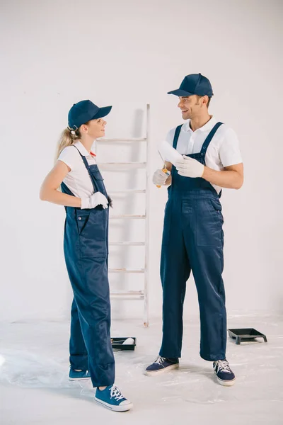 Pintor guapo en uniforme sosteniendo rodillo de pintura mientras habla con bastante colega - foto de stock