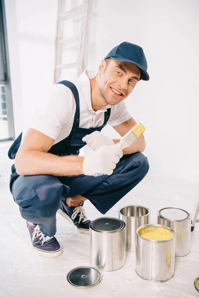 Handsome young painter in uniform holding paintbrush near cans with paint and smiling at camera — Stock Photo