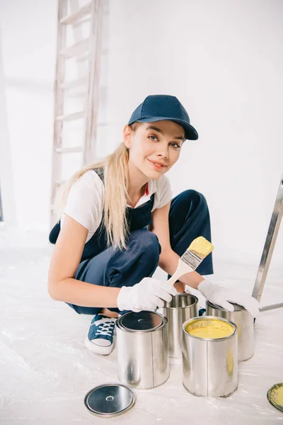 Belo jovem pintor em macacão e boné segurando pincel perto de lata com tinta amarela — Fotografia de Stock