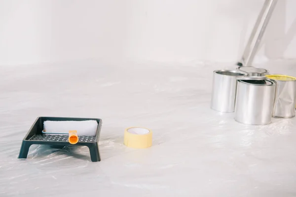 Boîtes de conserve avec peinture, rouleau de peinture dans un bac à rouleaux et ruban adhésif sur sol blanc recouvert de cellophane — Photo de stock