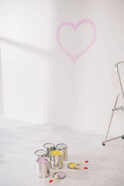 Quarto com coração rosa desenhado na parede branca, latas com tinta e pincéis no piso branco coberto com celofane — Fotografia de Stock