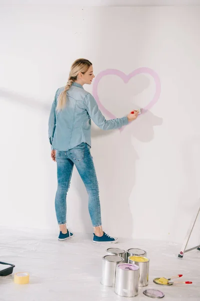 Pretty young woman drawing heart with pink paint on white wall — Stock Photo