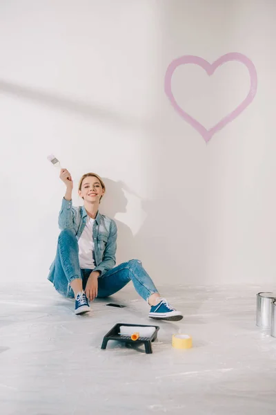 Feliz joven sentada debajo de la pared blanca con pintado de rosa cerca y sonriendo a la cámara - foto de stock