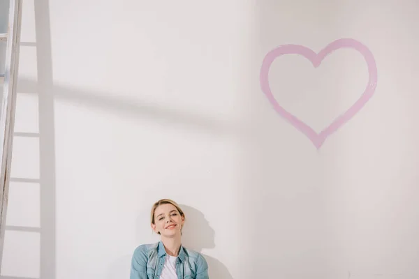 Happy young woman sitting on floor near white wall with painted pink heart — Stock Photo