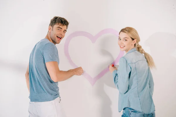 Feliz joven pareja sonriendo a la cámara, mientras que de pie cerca de corazón rosa dibujado en la pared blanca - foto de stock