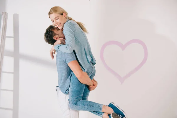 Heureux jeune homme tenant petite amie sur les mains près de coeur rose dessiné sur mur blanc — Photo de stock