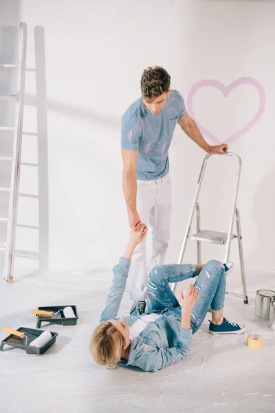 Young man helping girlfriend fallen from ladder and lying on floor — Stock Photo