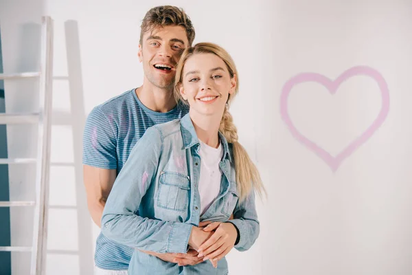 Happy young couple embracing and looking at camera while standing near white wall with drawn pink heart — Stock Photo