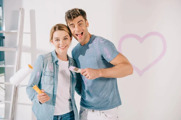 Heureux jeune couple regardant caméra tout en tenant pinceau rose et rouleau de peinture — Photo de stock