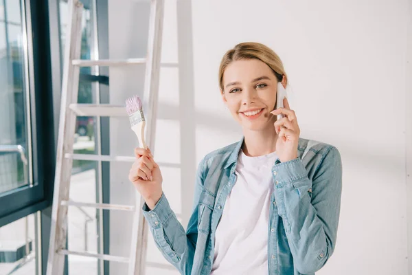 Atractiva joven que habla en el teléfono inteligente mientras sostiene el pincel rosa y sonríe a la cámara - foto de stock