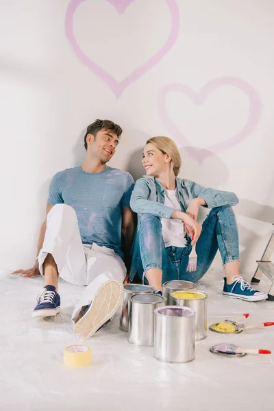 Feliz joven pareja sentada en el suelo por la pared blanca bajo dibujado corazones rosados - foto de stock