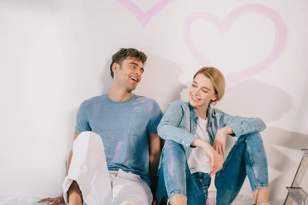 Счастливая молодая пара сидит у белой стены под нарисованным розовым сердцем — стоковое фото