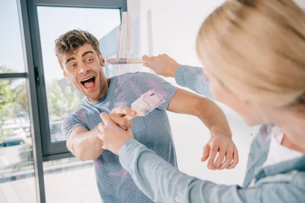 Alegre joven pareja divertirse y luchar en rosa pinceles mientras que la reparación de la habitación - foto de stock