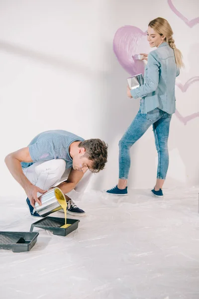 Young man adding yellow paint into roller tray while girlfriend drawing pink heart on white wall — Stock Photo