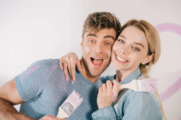 Glückliches junges Paar umarmt und lächelt in die Kamera, während es rosa Pinsel in der Hand hält — Stockfoto