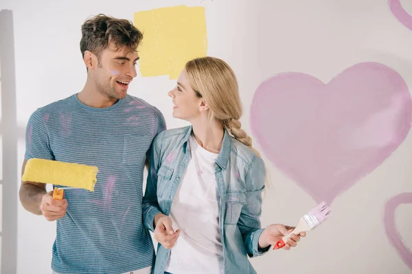 Feliz joven pareja mirando el uno al otro mientras que de pie cerca de corazón rosa pintado en la pared blanca - foto de stock