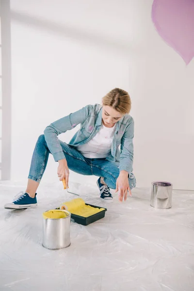 Atractiva joven mujer poniendo rodillo de pintura en la bandeja de rodillos con pintura amarilla - foto de stock